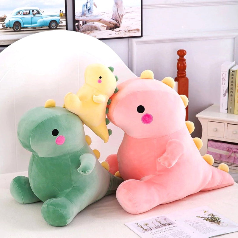 Three adorable dinosaur plush toys in soft fabric, featuring a large pink dino, a small yellow dino, and a large green dino, displayed on a cozy bed with decorative elements in the background.